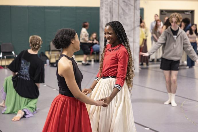 Two students in long skirts smile at each other as they hold hands.